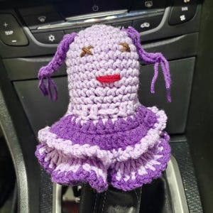 May include: Purple crocheted doll with a ruffled skirt and a smiling face. The doll has two pigtails and is sitting in a car.