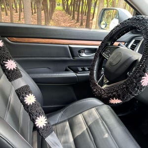 May include: Black crochet car seat belt covers and steering wheel cover with pink flower accents. The covers are designed to fit a car's interior and add a decorative touch. The steering wheel cover is shown on a black steering wheel.