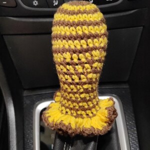 Puede incluir: Funda de palanca de cambios de coche hecha a mano, tejida a ganchillo. La funda es amarilla y marrón, con un patrón de rayas en el cuerpo principal y un borde con volantes en la base. Diseñada para la palanca de cambios de un coche.