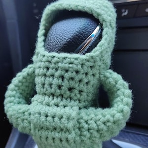 May include: A green crocheted  gear shift cover shaped like a person with arms crossed. The cover is made of soft yarn and has a  hooded head.