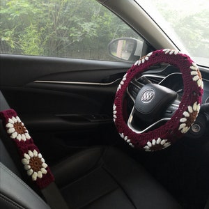 May include: A burgundy crocheted steering wheel cover with white daisy-like flowers. The cover is fitted to a black leather steering wheel in a car.