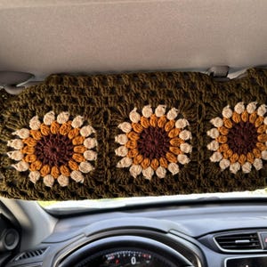 Op de afbeelding: Een handgemaakte, olijfgroene gehaakte zonneklep met drie zonnebloemontwerpen. De zonnebloemen hebben bruine, oranje en crèmekleurige bloemblaadjes. De zonneklep is in een auto geïnstalleerd, met het dashboard en de voorruit op de achtergrond.