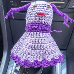 May include: A crocheted purple and gray doll with a purple dress and pigtails. The doll is sitting on a car's gear shift.