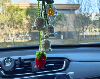 Op de afbeelding: Een gehaakte autohanger met een bloem en een aardbei die aan een achteruitkijkspiegel hangt. De hanger heeft witte klokvormige bloemen, een gele madeliefje en een rode aardbei.