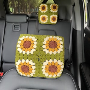 May include: Two-piece set of crochet car accessories. The set includes a headrest cover and a seat cushion, both featuring a sunflower design. The sunflowers have brown, yellow, and cream-colored petals on a green background. The items are in a car.