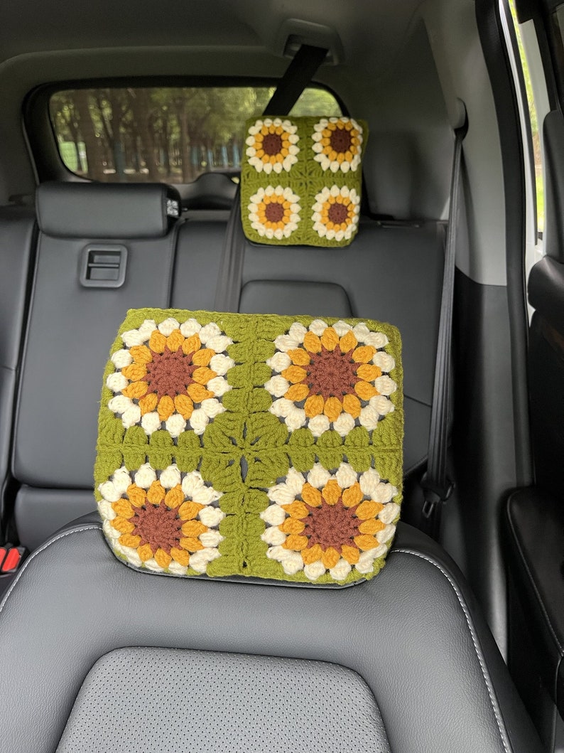 May include: Two-piece set of crochet car accessories. The set includes a headrest cover and a seat cushion, both featuring a sunflower design. The sunflowers have brown, yellow, and cream-colored petals on a green background. The items are in a car.