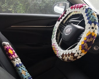 May include: A colorful crocheted steering wheel cover with a white background and brown, pink, yellow, green, and purple accents. The cover is on a black leather steering wheel in a car.