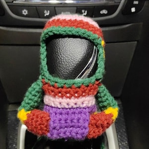 Op de afbeelding: Een kleurrijke gehaakte versnellingspookhoes in de vorm van een persoon. De hoes is gemaakt van groen, rood, roze, geel en paars garen. De hoes zit op een zwarte versnellingspook in een auto.