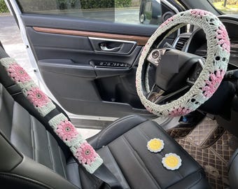 May include: A car interior with crocheted seatbelt covers and a crocheted steering wheel cover. The covers are pink and white with a floral design. There are also two yellow crocheted flowers on the seat.