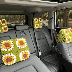May include: Crocheted car headrest covers featuring a sunflower design. The covers are in shades of green, yellow, brown, and white. The headrests are installed in a car with black leather seats. The background shows a view of trees.