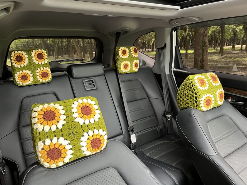 May include: Crocheted car headrest covers featuring a sunflower design. The covers are in shades of green, yellow, brown, and white. The headrests are installed in a car with black leather seats. The background shows a view of trees.