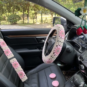 K&ouml;nnte beinhalten: Ein Autoinnenraum mit geh&auml;kelten rosa und wei&szlig;en, blumenf&ouml;rmigen Sicherheitsgurt- und Lenkradbez&uuml;gen. Das Auto hat eine schwarze Lederausstattung und ein schwarzes Armaturenbrett.