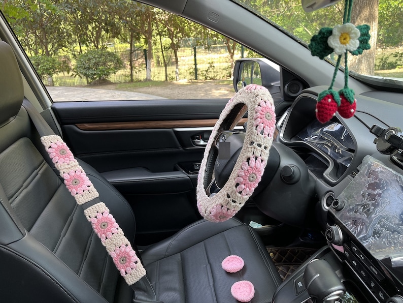 K&ouml;nnte beinhalten: Ein Autoinnenraum mit geh&auml;kelten rosa und wei&szlig;en, blumenf&ouml;rmigen Sicherheitsgurt- und Lenkradbez&uuml;gen. Das Auto hat eine schwarze Lederausstattung und ein schwarzes Armaturenbrett.