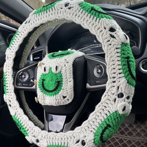 May include: A white and green crocheted steering wheel cover with a smiley face design. The cover is made of yarn and has a soft texture.