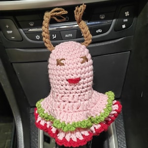 May include: A pink crocheted car gear shift cover with green and red trim. The cover is shaped like a person with brown yarn hair and a smiling face.