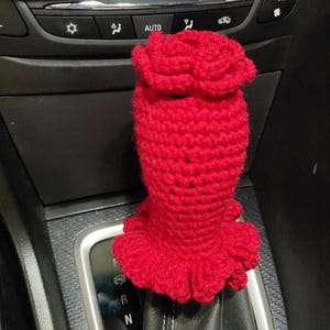 Könnte beinhalten: Ein roter, gehäkelter, rosenförmiger Schaltknauf mit einem gerüschten Boden. Der Knauf ist am Schalthebel eines Autos befestigt.
