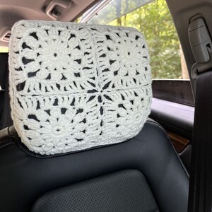 May include: White crocheted headrest for a car seat. The headrest is made with a square pattern and has a flower design in the centre of each square.