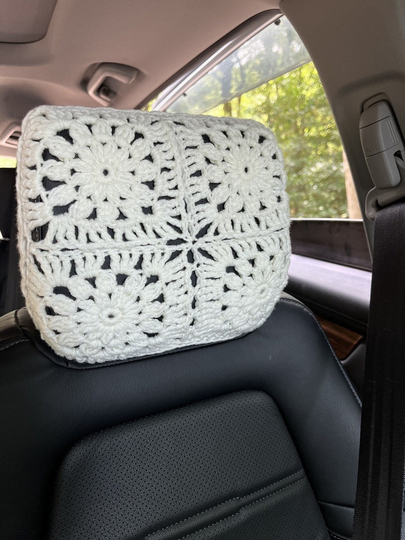 May include: White crocheted headrest for a car seat. The headrest is made with a square pattern and has a flower design in the centre of each square.