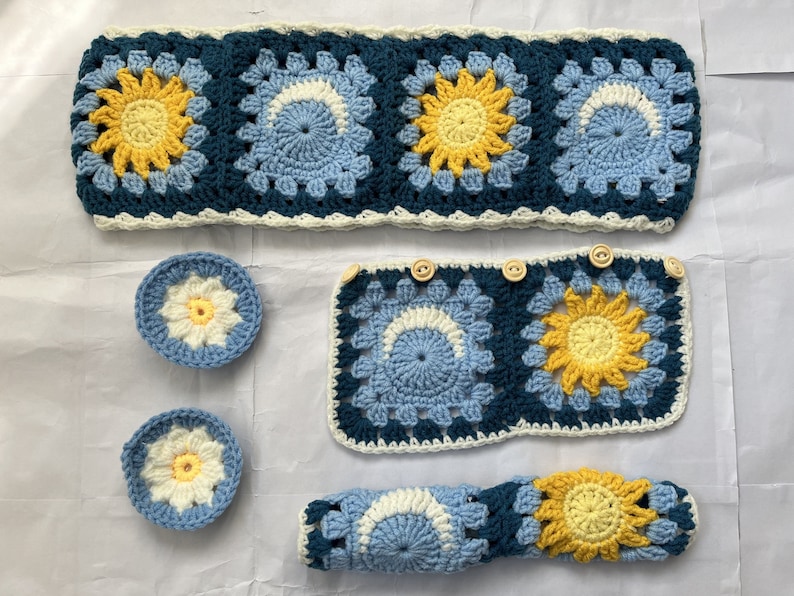 May include: A set of crocheted items in blue, white, and yellow. The set includes a headband, a square, two small circles, and a long rectangle. The items are all made with a granny square stitch and feature a sunflower design.