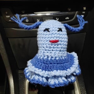 May include: A blue crocheted doll with a red mouth and blue yarn hair. The doll is wearing a blue and light blue ruffled skirt. The doll is sitting on a car's gear shift.