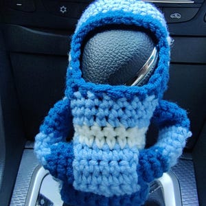 May include: A blue and white crocheted car gear shift cover. The cover is shaped like a person with a round head and arms folded across the chest.