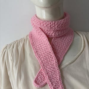 May include: A light pink crochet scarf displayed on a mannequin. The scarf is handmade with a textured stitch, featuring a neck warmer and long, crossed ends. The soft pink color adds a touch of warmth and style.
