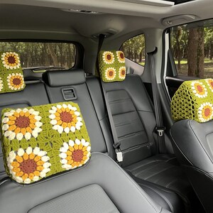 May include: Crocheted car headrest covers featuring a sunflower design. The covers are square, with a green background and four sunflower motifs in yellow, brown, and white. The headrests are placed on black car seats.