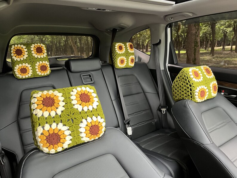 May include: Crocheted car headrest covers featuring a sunflower design. The covers are square, with a green background and four sunflower motifs in yellow, brown, and white. The headrests are placed on black car seats.
