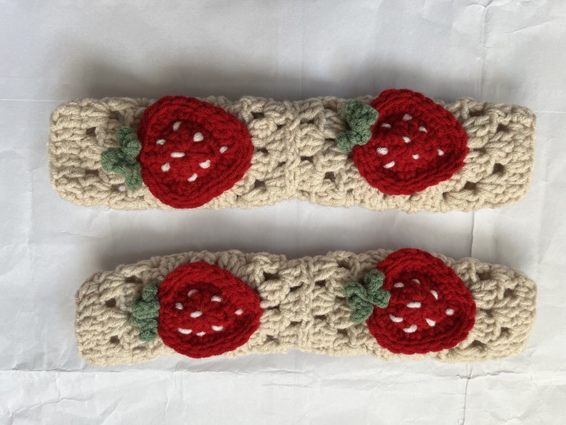 May include: Two beige crocheted headbands with red and green strawberry appliques. Each headband has a single strawberry with white stitching for the seeds.