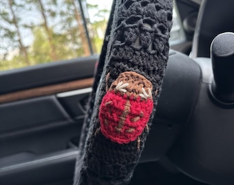 May include: Black crochet steering wheel cover with red and brown ladybug accents. The cover is designed to fit a standard steering wheel and features a unique, handmade design. The ladybugs are placed around the cover.