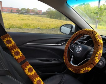 May include: A crocheted sunflower design car steering wheel cover and matching seatbelt cover. The steering wheel cover is brown with yellow sunflowers and the seatbelt cover is brown with yellow sunflowers.