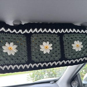 May include: Hand-crocheted car sun visor with a daisy pattern. The black visor has a white zig-zag trim and three green squares, each with a white daisy and yellow centre. A decorative, handmade car accessory.