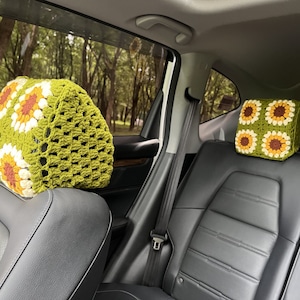 May include: Two crochet car headrest covers. The covers are green with a sunflower pattern in white, yellow, and brown. The headrests are installed in a car with a black leather interior. The background shows a view of trees.