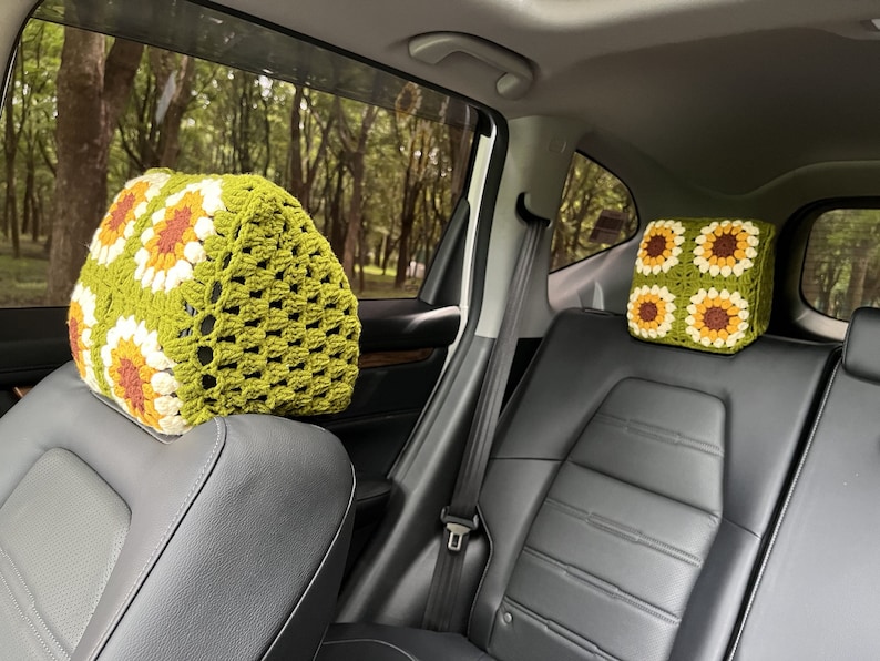 May include: Two crochet car headrest covers. The covers are green with a sunflower pattern in white, yellow, and brown. The headrests are installed in a car with a black leather interior. The background shows a view of trees.