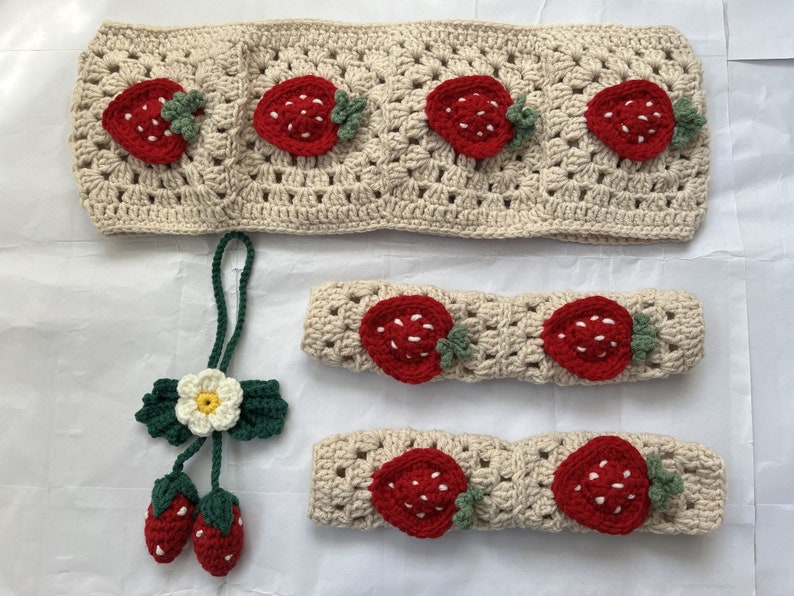 May include: A set of crocheted strawberry-themed headbands and a matching keychain. The headbands are beige with red and green strawberries. The keychain has a green stem, a red strawberry, and a white flower.
