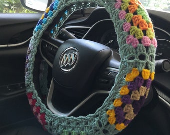 May include: A colorful, crocheted steering wheel cover. The cover features a granny square design with various colors, including green, pink, yellow, purple, and brown. The cover is on a black steering wheel inside a car.