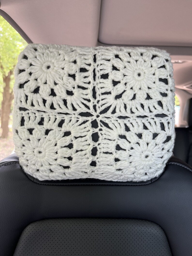May include: White crocheted headrest cover with a floral pattern. The cover is made of a soft, white yarn and is perfect for adding a touch of elegance to any car interior.
