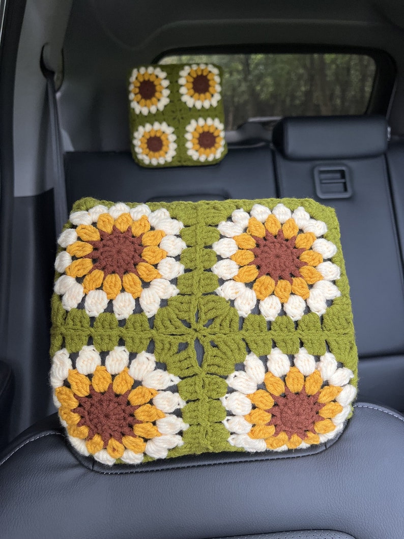 May include: Crocheted car headrest covers featuring a sunflower design. The covers are made with green, brown, yellow, and cream-colored yarn. The sunflower pattern is centered on a green square. The headrest covers are placed on a black car seat.