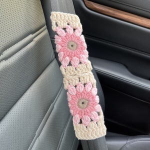 K&ouml;nnte beinhalten: Ein geh&auml;kelter Sicherheitsgurtbezug mit rosa und cremefarbenen Blumen. Der Bezug ist aus Garn und hat eine quadratische Form.