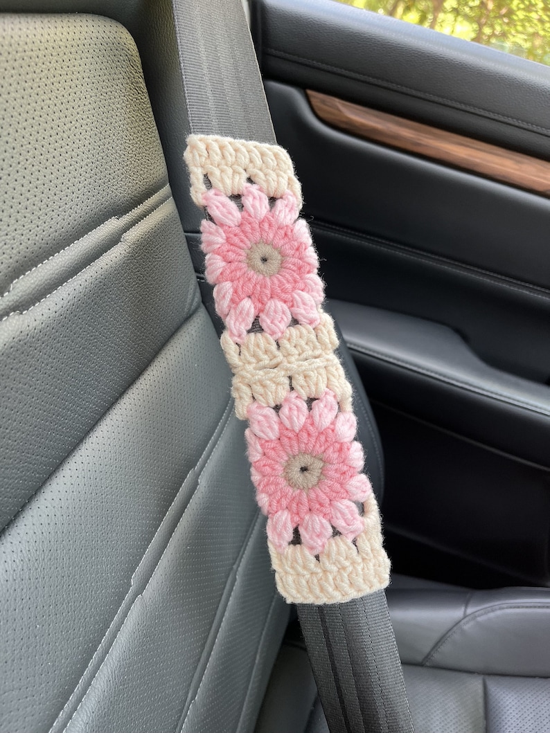 K&ouml;nnte beinhalten: Ein geh&auml;kelter Sicherheitsgurtbezug mit rosa und cremefarbenen Blumen. Der Bezug ist aus Garn und hat eine quadratische Form.