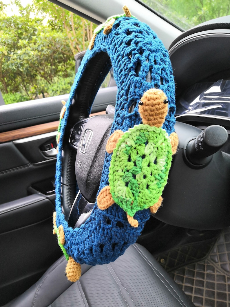 May include: A blue crocheted steering wheel cover with two green and brown crocheted turtles attached. The cover is in a car.