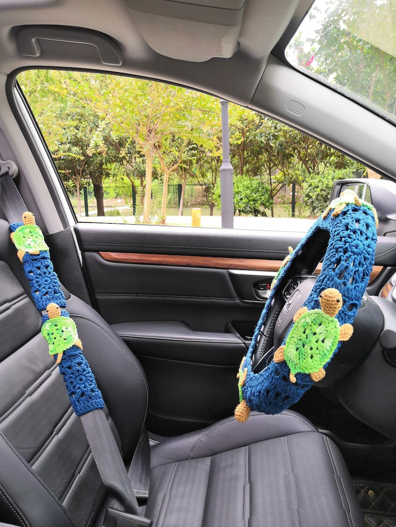 May include: A blue and green crocheted steering wheel cover with a turtle design. A matching blue and green crocheted seatbelt cover with a turtle design is also visible.