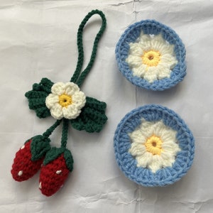 May include: A set of three crocheted items: a flower with green leaves, two red strawberries, and two blue and white daisy-shaped coasters.