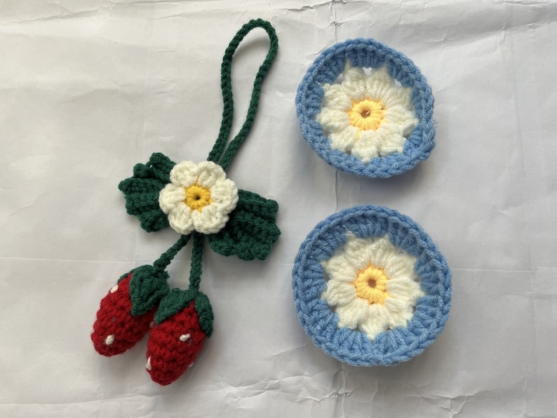 May include: A set of three crocheted items: a flower with green leaves, two red strawberries, and two blue and white daisy-shaped coasters.
