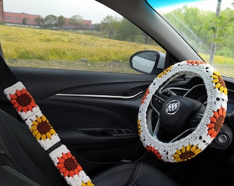 May include: A crocheted steering wheel cover with white, brown, and orange flowers. The cover is on a black leather steering wheel in a car.