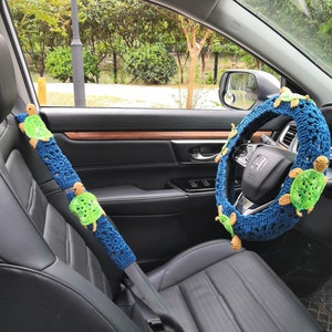 May include: A blue and green crocheted steering wheel cover with a turtle design. The cover is fitted to a black leather steering wheel in a car.