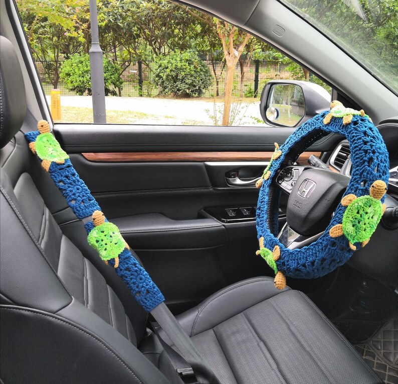 May include: A blue and green crocheted steering wheel cover with a turtle design. The cover is fitted to a black leather steering wheel in a car.