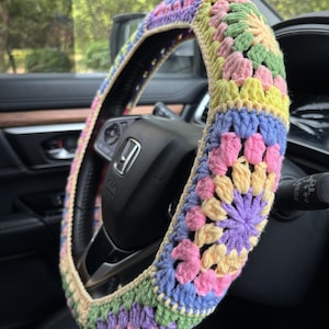 May include: A colorful, crocheted steering wheel cover. The cover features a patchwork design with pastel pink, yellow, blue, green, and purple floral patterns. The cover is installed on a black steering wheel inside a car.