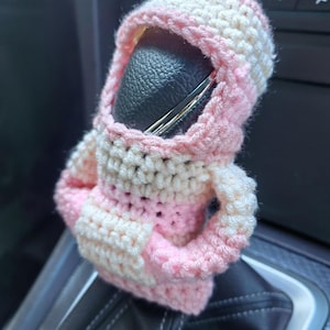 May include: A pink and white crocheted car gear shift cover. The cover is shaped like a person with a hood.
