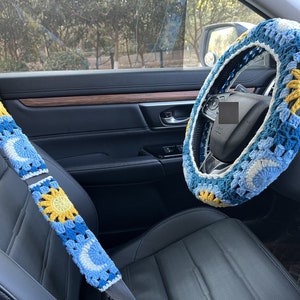May include: A blue and yellow crocheted steering wheel cover with a sun and moon design. The cover is on a black leather steering wheel in a car.